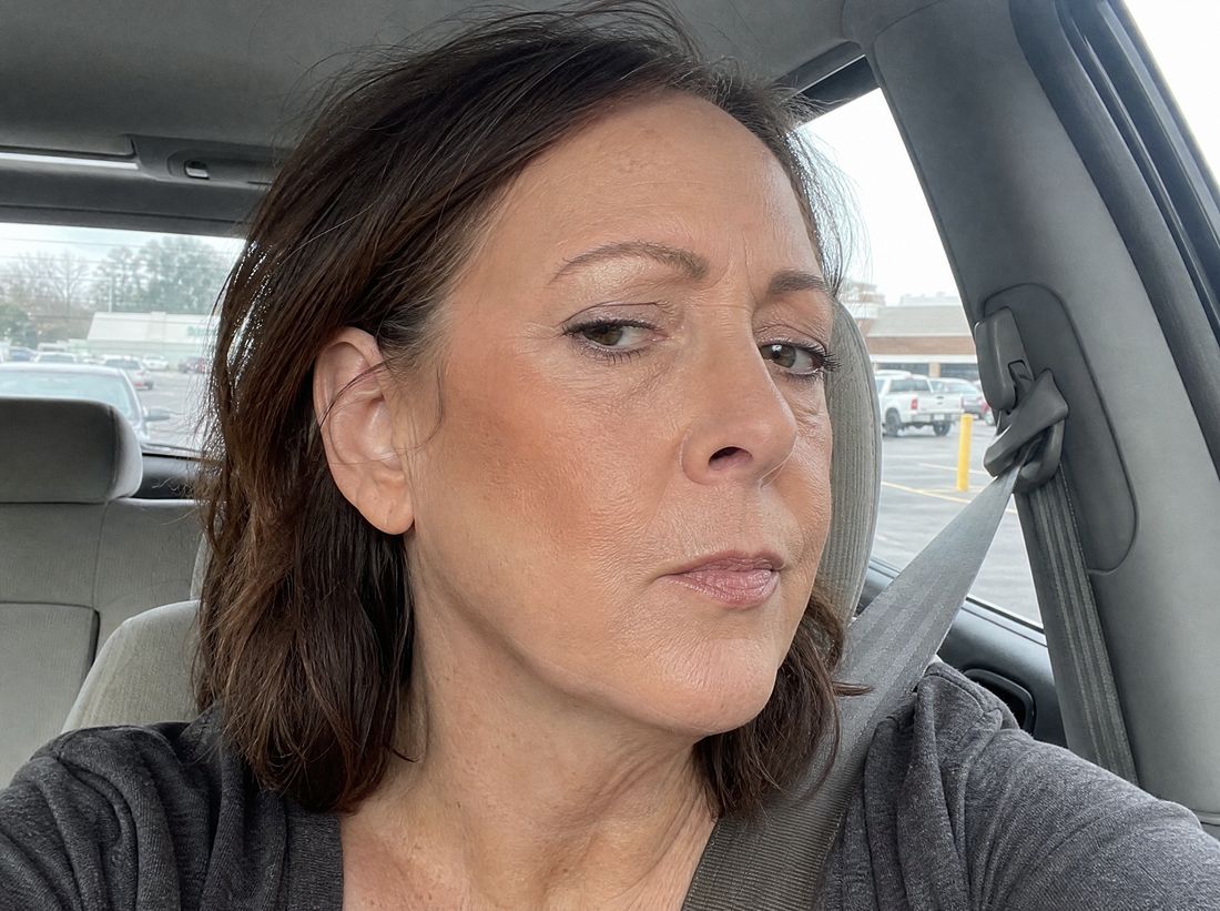 A woman catching her reflection in the car mirror in daylight, foundation reading warm against her neck